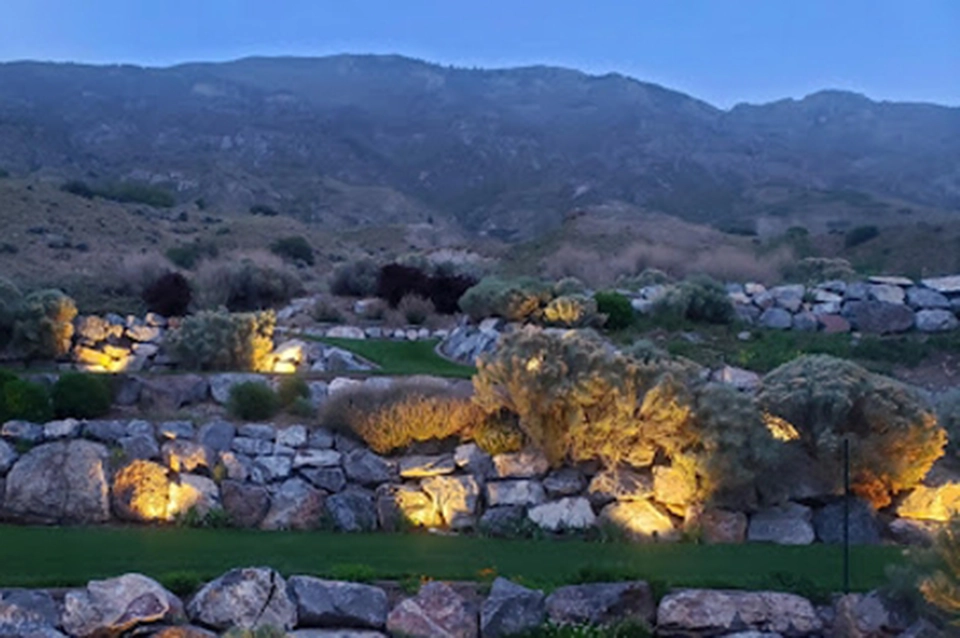 Rock terraces with shrubs and small bushes are softly illuminated by ground lights at dusk, set against a backdrop of hills and a cloudy blue sky.