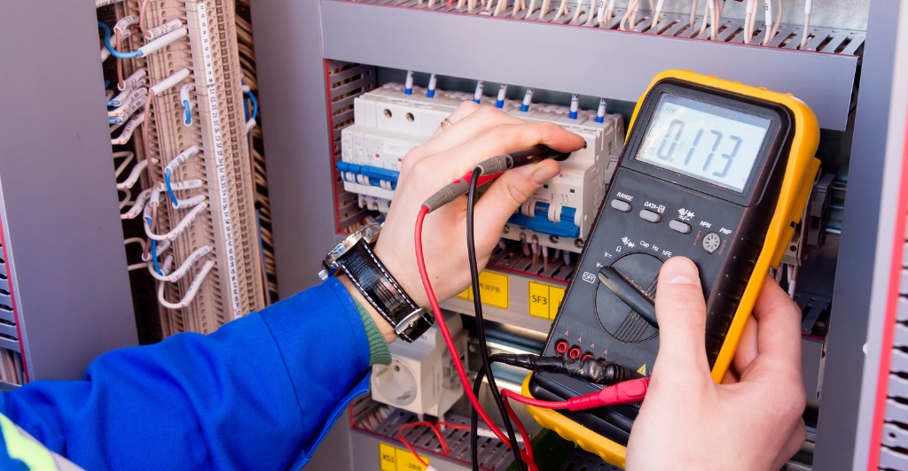 A person uses a digital multimeter to measure voltage in an electrical control panel with multiple wires, switches, and circuit breakers.