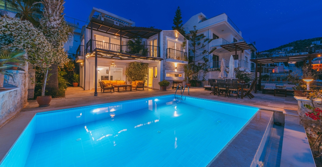 A brightly lit swimming pool in front of a modern villa at night, with outdoor seating on a patio, surrounded by plants and neighboring buildings under a deep blue sky.